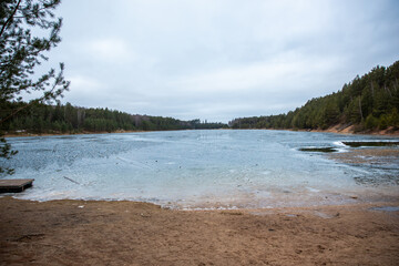 A partially frozen lake with sandy shores in the foreground, dense evergreen forests on both sides, and a small wooden dock on the left under an overcast sky.
