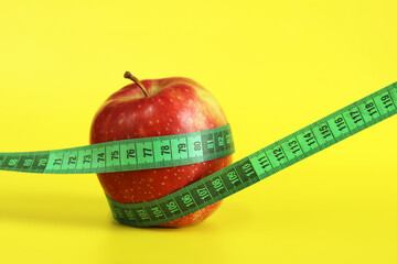 Measuring tape wrapped around an apple. Metaphor - round figure. Ripe red apple and tape measure on yellow background. Concept of healthy eating, diet and weight loss. Body volume control