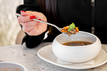 A close-up of a person's hand holding a spoonful of soup with mushrooms and vegetables. The soup is...
