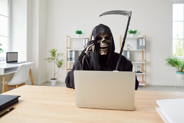 Business man wearing Halloween grim reaper death costume holding scythe sitting at the desk on his...