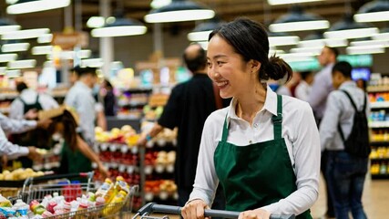 A cheerful worker in a green apron pushes a cart in a bustling supermarket. The video captures a lively, candid moment from a medium angle.