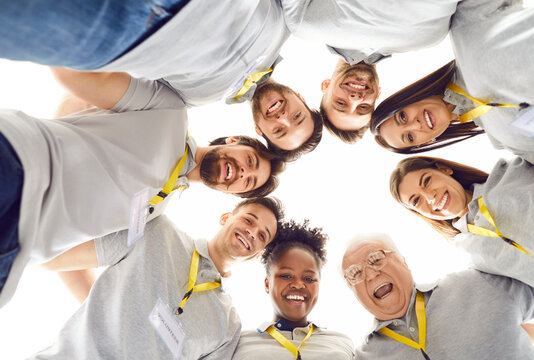 Team of happy diverse volunteers and friends having fun together. From below low angle view group of cheerful smiling multiracial young and elderly people in uniforms huddling looking down at camera