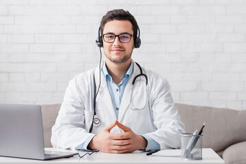 Personal blog of doctor. Smiling man in white coat with headphones, sits at table with laptop in living room interior, free space