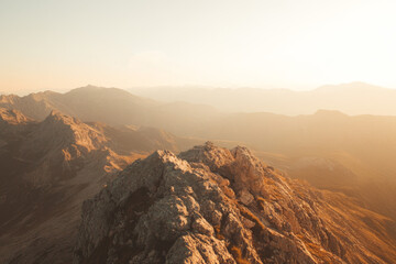 A breathtaking mountainous landscape during sunrise or sunset, with warm golden light casting a serene and dramatic atmosphere over the rugged terrain and distant peaks. Subtle grain, soft focus edges