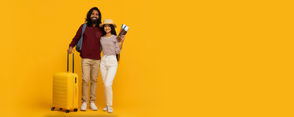 Happy traveling couple standing close with passports and a yellow suitcase on a sunny yellow background, copy space