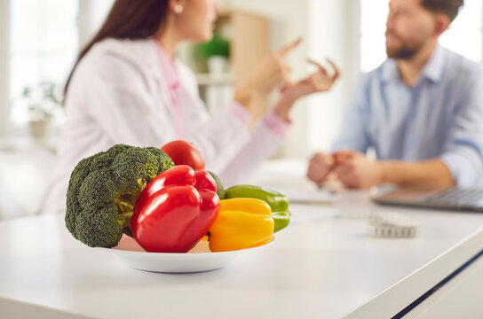 Fresh healthy vegetables on a plate in the foreground in nutritionist doctor's office. Dietician woman sitting on workplace and giving consultation to man patient. Healthy nutrition concept.