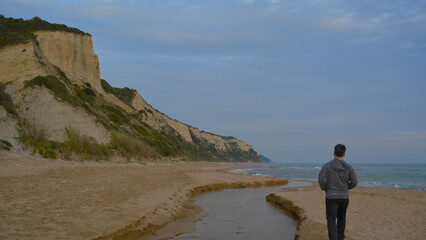 Man walking on the beach with rocky mountains and  cloudy sky as night falls. Sunset on Corfu Island, cool weather.