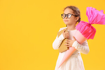 Happy little girl in eyeglasses with pink school cone and book on yellow background