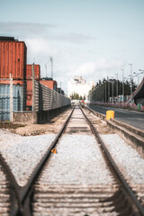 Obraz premium Vertical telephoto shot of industrial railway track leading towards a distant grain silo, flanked by shipping containers and a fenced-off storage area. Parallel city street lined with lampposts