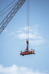 A heavy-duty industrial crane lifting a red steel spreader bar with multiple latches and mechanisms...