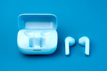 A pair of modern wireless earbuds placed next to an open charging case on a blue background. A...