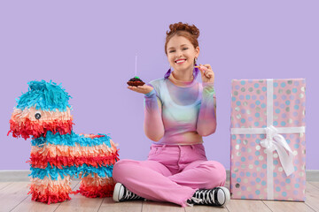 Beautiful young happy woman with gift box, pinata and birthday cupcake sitting near purple wall