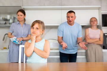 At home kitchen, parents scold daughter for careless act. Sulky preschool girl stand near table during serious conversation with parents, mom clean dish in background
