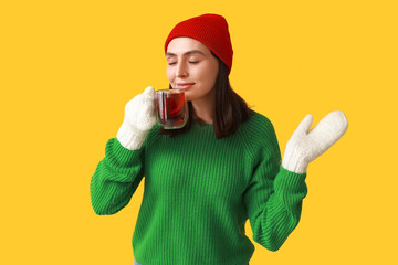 Beautiful young woman with glass cup of hot mulled wine on yellow background