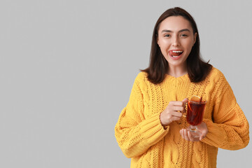 Funny young woman with glass cup of hot mulled wine on grey background