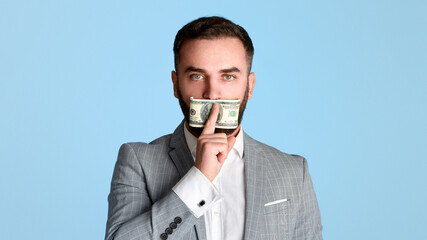 Silenced by money concept. Young businessman covering his mouth with hundred dollar bill over blue...