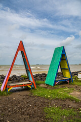 two benches in the shape of an isosceles triangle with a pointed top on the beach, the benches face the sea with a clear sky in the background for advertising space.