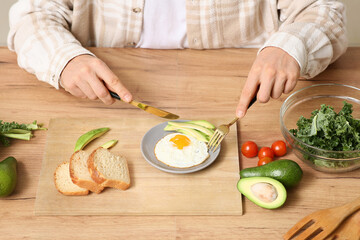 Young man eating tasty fried egg with fresh avocado, bread and kale salad at wooden table