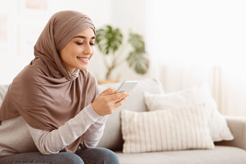 Online Banking. Smiling Muslim Woman In Hijab Using App On Smartphone At Home, Sitting On Couch In...