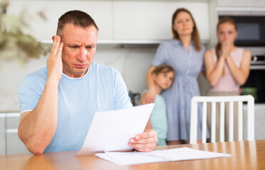 Upset adult man sitting at kitchen table at home and looking at papers anxiously. Worried wife and two teenage daughters standing behind on blurred background