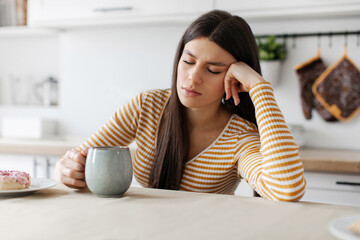 Tired woman with coffee cup sleeping on table in kitchen interior, sitting with closed eyes....