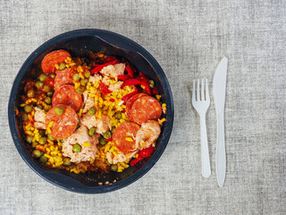 Spanish paella dish in black round plastic tray on light color table cloth and plastic forks and knife. Popular food with rice and sea products and meat. Fast food product.