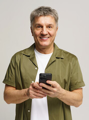 Smiling older man in casual clothing holding smartphone and standing against light backdrop