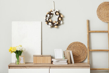 Chest of drawers with tulips, books and Easter wreath on light wall in room