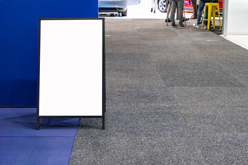 a blank white empty mockup of sandwich board or A-frame sign with copy space for customize, in an exhibition hall, trade show, or commercial space with people or visitors standing in the background. 