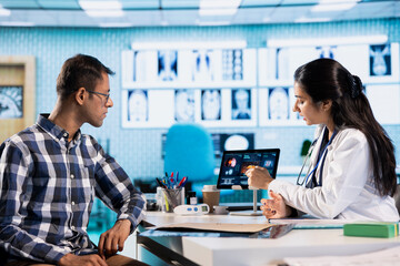 Indian expert explaining diagnostics through test results with a male patient at medical office, doing scan interpretation at check up with young man. Healthcare services on insurance.