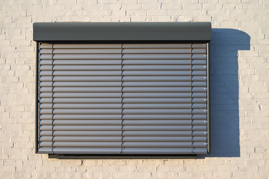 Modern window with closed horizontal metal roller shutters installed on a white brick wall