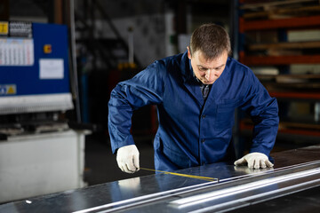 Adult man in uniform measures sheet of metal with tape measure at metallurgical factory