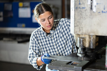 Focused young female worker operating hydraulic punch press in manufacturing facility, creating precise holes in metal sheets..