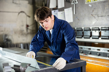 Guy working at metallurgical plant measures sheet of iron using tape measure and marks the cutting line