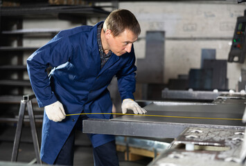 Male worker at metallurgical plant measures an iron sheet using tape measure and marks the cutting...