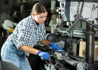 Young woman in casual plaid shirt and jeans equipped with protective gloves, focusing on metalworking in professional workshop, confidently using drill press to create holes in workpiece