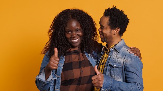Affectionate husband and wife giving a like symbol on camera, african american couple posing with thumbs up in studio. Married people giving their approval and nodding in agreement. Camera A.