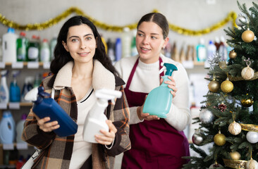 Polite young saleswoman offering spray cleaner to female customer in store decorated for Xmas