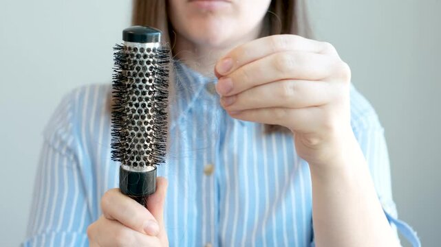 Portrait of a young woman taking out fallen hair from a round comb front view close-up on a gray background.