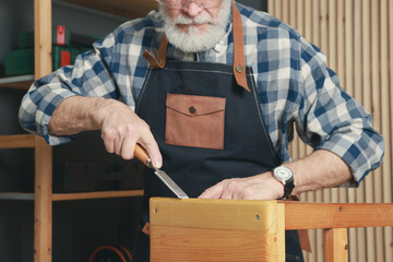 Relaxing hobby. Senior man repairing wooden stool with chisel in workshop, closeup