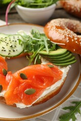 Delicious bagel with salmon, cream cheese, cucumber, avocado and pumpkin seeds on table, closeup
