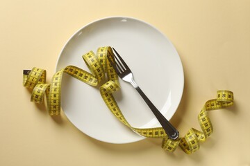 Bulimia. Plate with fork and measuring tape on beige background, top view