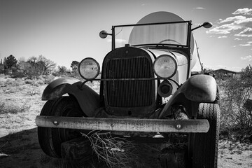 An antique Model A truck hauling a large tank in the desert. Black and white.