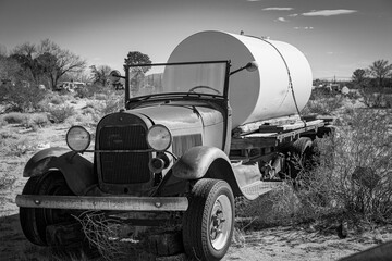 An antique Model A truck hauling a large tank in the desert. Black and white.