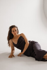 Fototapeta premium A young Caucasian woman with long brown hair poses gracefully on a white backdrop. She wears a dark slip dress, exuding elegance and confidence. Concepts of beauty, fashion, and femininity.