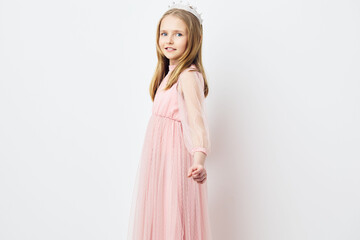 Cute girl in a pink dress with a tiara, smiling gently, stands against a simple white background,...