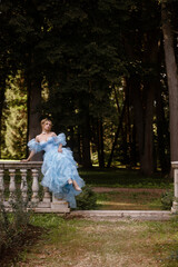 Graceful young woman in elegant light blue dress posing near stone balustrade in picturesque garden. Concept fantasy or historical themes.