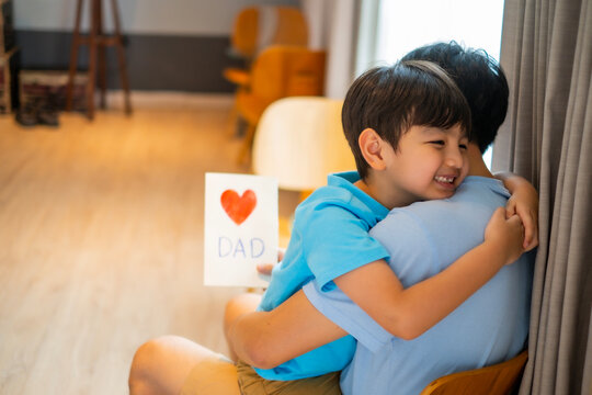Asian little child give a card with heart drawing and word dad with to his father for surprising, Dad and son embrass each other with love at livining room. Father's day concept.