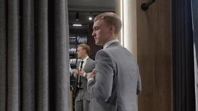 Elegant businessman adjusting his suit jacket while looking in the mirror in the dressing room of a menswear store, choosing his outfit for a special occasion or business meeting