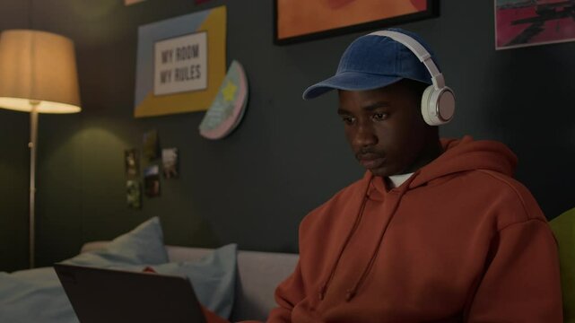 Modern African American boy sitting on sofa in dorm room, listening to music in headphones and typing on laptop